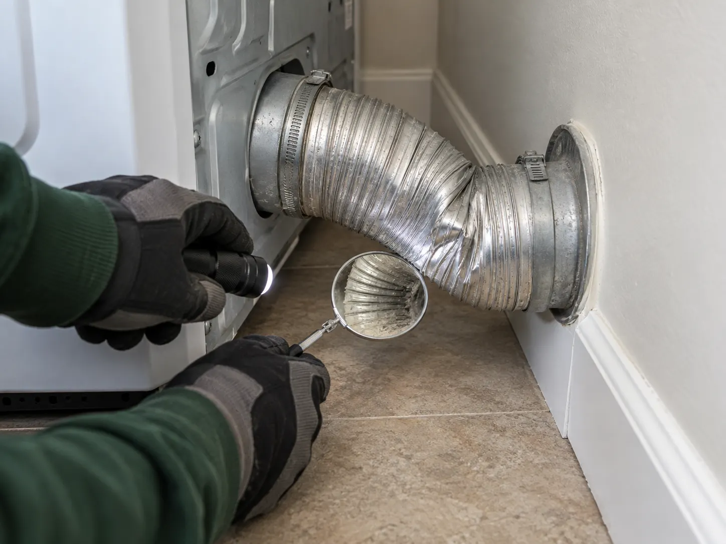 Technician using a flashlight and inspection mirror to examine a sharply bent dryer exhaust connector behind the appliance.