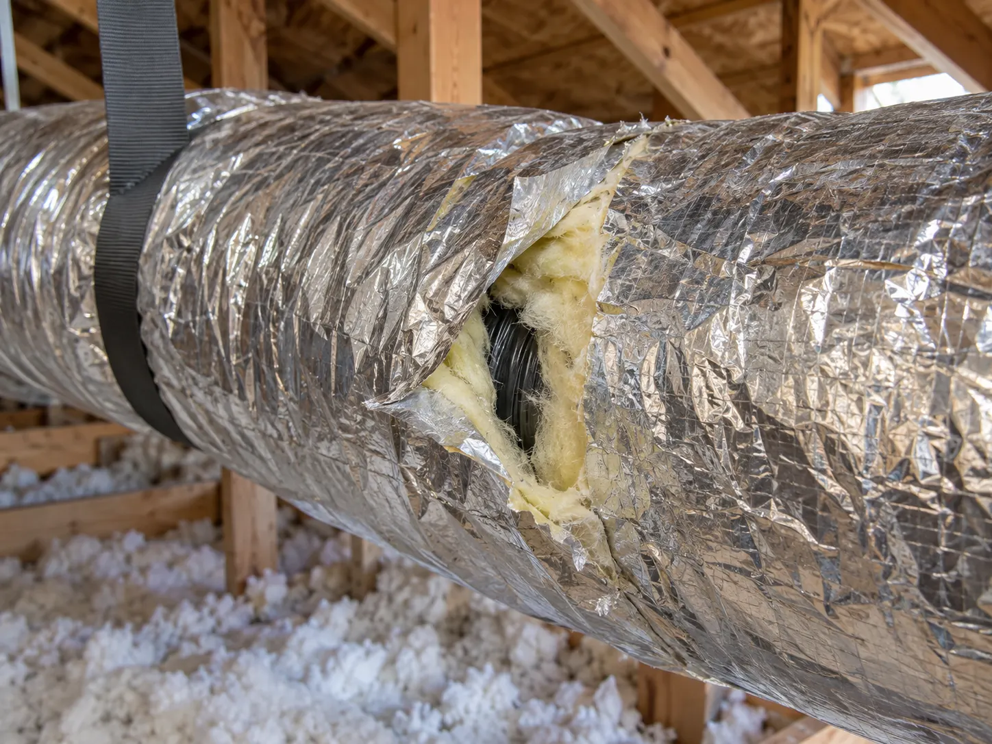 Damaged outer duct covering exposing fiberglass insulation beneath the silver jacket in an attic.